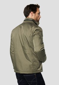 Olivově zelená prošívaná bunda se stojáčkem, zapínáním na zip a logem na rameni, ve spojení s tmavě modrými džínami.