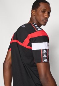 Μαύρο κοντομάνικο μπλουζάκι με κόκκινες και λευκές λεπτομέρειες. Λογότυπο Kappa στον ώμο και τα μανίκια, με άνετη υφή από βαμβάκι.