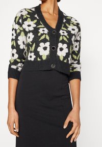 Gekürzter schwarzer Cardigan mit floralem Muster, der weiße Blumen und grüne Blätter zeigt, gestrickte Textur und drei metallische Knöpfe auf der Vorderseite hat.