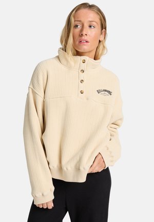 Beige ribbad tröja med hög krage, treknäppning och broderad logotyp. Manschetter med sömdetaljer. Mjuk textur.
