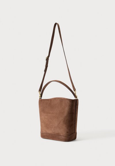 Sac seau en daim marron avec une poignée courte et une longue bandoulière réglable sur un fond uni.