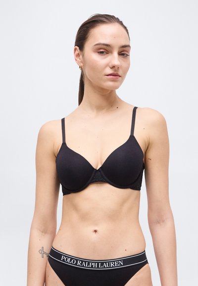 Femme aux cheveux bruns portant un soutien-gorge noir et des sous-vêtements Polo Ralph Lauren noirs, sur un fond blanc uni.