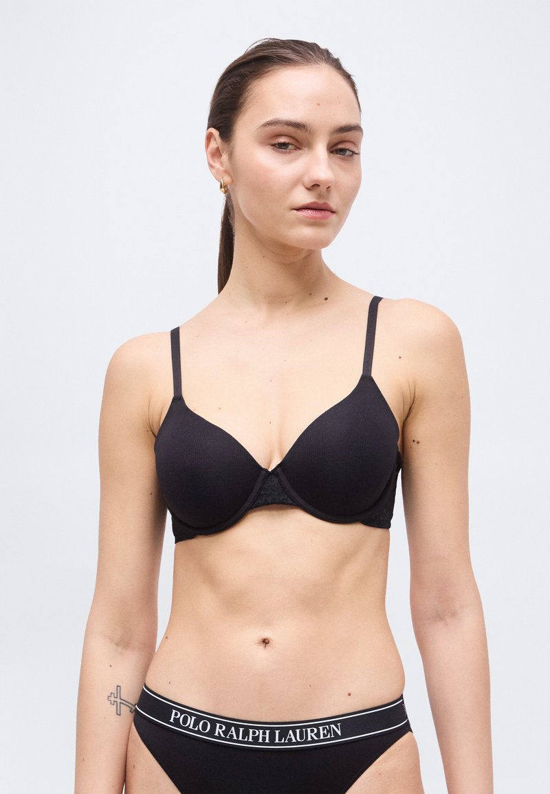 Γυναίκα με καστανά μαλλιά, φορώντας μαύρο σουτιέν και μαύρα εσώρουχα Polo Ralph Lauren, σε λευκό φόντο.