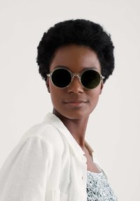 Pyöreät aurinkolasit tummilla linsseillä ja kirkkailla kehyksillä, päällä valkoinen paita, jossa on teksturoitu sininen kuviollinen yläosa, ja lyhyet kiharat hiukset.