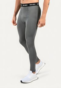 Homme portant un legging de sport ajusté gris avec une ceinture noire et des chaussures de course blanches sur un fond uni.