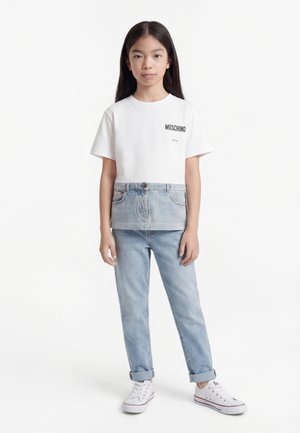 Enfant debout de face portant un T-shirt blanc Moschino avec un jean en denim et des baskets blanches, sur fond blanc uni.