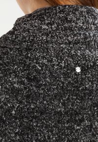 Strukturiertes schwarz-graues Strickgewebe mit einer weichen, flauschigen Oberfläche; hat eine silberne Logo-Knopf-Akzentuierung auf der Rückseite.