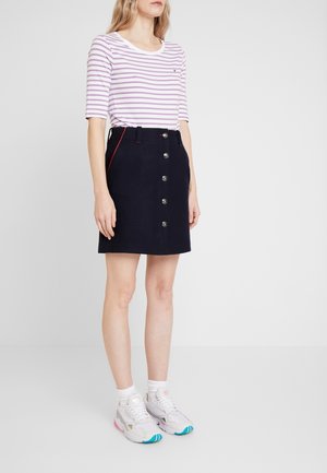 Jupe bleue marine avec des boutons sur le devant, des coutures rouges contrastantes, associée à un haut rayé violet et blanc, et des baskets blanches avec des accents colorés.