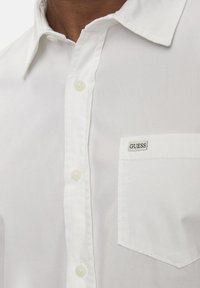 Witte button-up blouse van katoen, met een puntkraag, voorzak en klein "GUESS" logo op de zak. Zachte textuur.