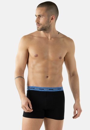 Homme torse nu avec des cheveux courts et des tatouages, portant un boxer noir avec une ceinture bleue, regardant vers sa gauche sur un fond uni.
