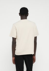 Μπεζ βαμβακερό t-shirt με στρογγυλή λαιμόκοψη και κοντά μανίκια, με άνετη εφαρμογή και μικρή ετικέτα της μάρκας στο κάτω αριστερό μέρος.