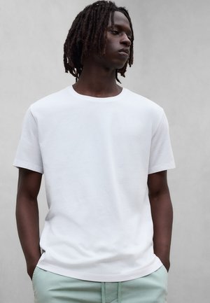 Ung mann med dreadlocks iført en ensfarget hvit kortermet T-skjorte og lys grønne bukser, hendene i lommer, ser til siden.