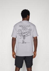 Siva bomba majica iz bombaža s grafičnim dizajnom koji prikazuje drvo i golf igrača. Natpisi "NIKE GOLF" i "Kada sumnjaš... 'Samo uradi to.'" u crnoj boji.