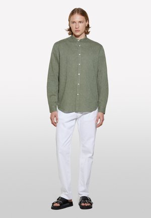 Chemise en lin vert à boutons avec un col montant, manches longues et ourlet droit, associée à un pantalon blanc et des sandales noires.