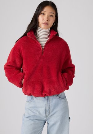 Femme portant un pull en polaire rouge avec une fermeture éclair quart de zip sur un col roulé rayé et un jean bleu clair, debout les mains dans les poches.