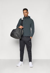 Nike-jakke i mørkegrå med hætte, bæret med sorte sweatpants og hvide sneakers. Modellen holder en sort duffelbag.