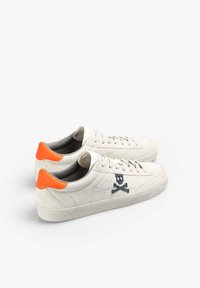 Beige Wildlederschuhe mit einer strukturierten Gummisohle, die ein orangefarbenes Fersenakzent und ein dunkelblaues Totenkopf- und Kreuzbones-Logo an der Seite aufweisen.