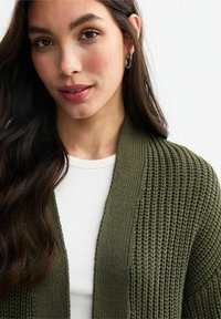 Knit cardigan in dark green with pronounced texture, open front design, and ribbed accents. Worn over a white top. Silver hoop earrings visible.