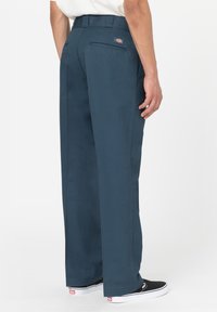 Muž v tmavě modrých pracovních kalhotách Dickies a černých slip-on teniskách Vans, stojící na bílém pozadí, boční pohled.