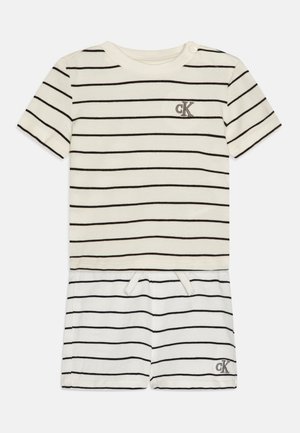 Csíkozott pamut kezeslábas krém és fekete színben. Rövid ujjú, kerek nyakú, középen dekoratív logóval. Állítható derék húzózsinórral.