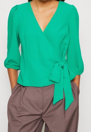 Zelený zavinovací top se tříčtvrtečními rukávy, texturovanou látkou a zavazovacím detailem v pase. Kombinován s volnými hnědými kalhotami.