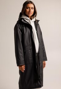 Manteau noir imperméable avec capuche, design long, fermeture éclair et boutons-pression, deux poches avant, superposé sur un sweat-shirt gris.