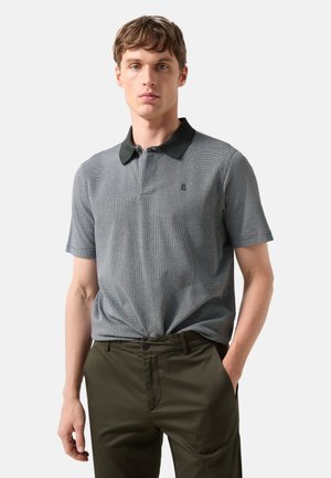 Młody mężczyzna ubrany w szarą koszulkę polo z krótkim rękawem i czarnym kołnierzykiem oraz ciemnozielone spodnie, stoi z jedną ręką w kieszeni na jednolitym tle.