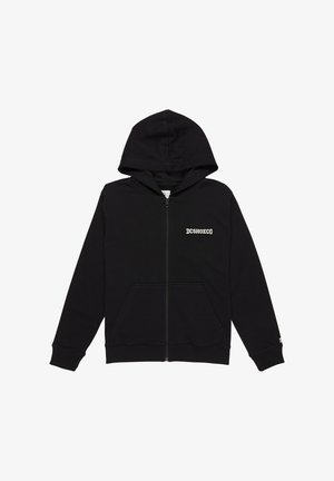 Blond chłopiec stoi z rękami w kieszeniach, ubrany w czarną bluzę z zamkiem, jasnoniebieskie dżinsy i czarno-pomarańczowe sneakersy na białym tle.