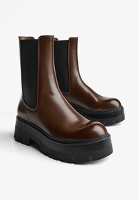 Cizme Chelsea din piele maro, cu panouri elastice negre pe lateral și tălpi groase de cauciuc negru, prezentând o textură netedă și un design cu vârf rotund.