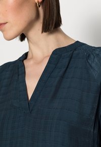 Blusa blu scuro con scollatura a V, con strisce testurizzate e dettagli arricciati sulle spalle. Tessuto morbido e vestibilità comoda.