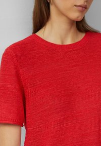 Rode gebreide trui met korte mouwen, geribbelde halslijn en een textuurafwerking. De stof lijkt zacht en lichtgewicht te zijn.