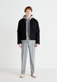 Black cropped jacket over a grey hoodie, paired with light grey trousers and white sneakers with red accents. Minimalist design and textures.