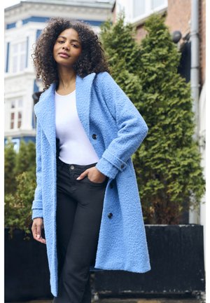 Manteau en laine bleu texturé avec de grands revers et deux boutons noirs, porté sur un haut blanc et un pantalon noir, sur fond de verdure et de bâtiments.
