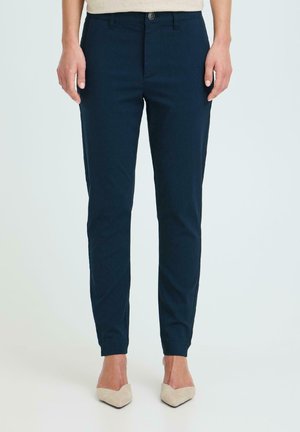 Femme portant un pantalon bleu marine ajusté et des ballerines beiges pointues, debout devant un fond clair uni.