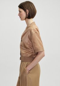 Kurze, braune Satinjacke mit V-Ausschnitt und geripptem Saum; das Model hat eine Hand in der Tasche und trägt ein goldenes, abstraktes Armreif.