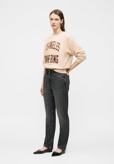 Kvinne som står iført beige genser med teksten «LOS ANGELES», svarte jeans, svart belte og svarte spisse sko mot hvit bakgrunn.