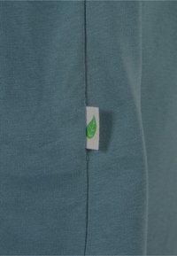Close-up van petrolkleurige stof met een klein wit label met een groen bladsymbool dat in een naad is genaaid.