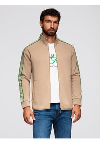 Giacca beige con zip e strisce verdi lungo le maniche. Tessuto leggero, due tasche laterali, colletto alto, design aderente.