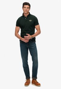 Donkergroen poloshirt met geborduurd logo, gecombineerd met blauwe jeans en bruine veterlaarzen. Strakke lijnen en een getailleerde pasvorm.