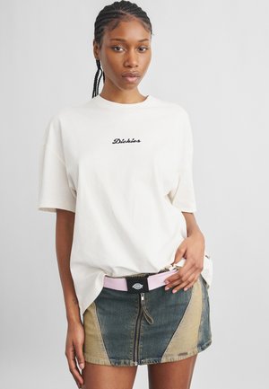 Jeune femme aux cheveux tressés portant un t-shirt blanc Dickies et une mini-jupe en denim bicolore avec une fermeture éclair à l'avant et une ceinture rose.