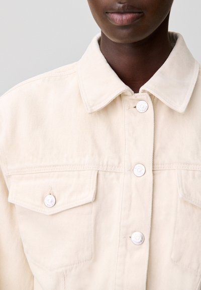 Chaqueta de mezclilla beige claro con cuello puntiagudo, dos bolsillos en el pecho y cierres con botones plateados. La textura es suave y ligeramente estructurada.