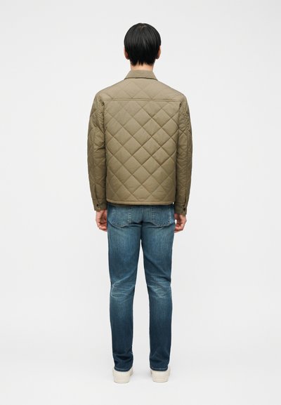 Άτομο με κοντά σκούρα μαλλιά στέκεται με την πλάτη προς τα εμπρός, φοράει μια φουσκωτή ελιά μπουφάν, μπλε τζιν και λευκά αθλητικά παπούτσια σε λευκό φόντο.