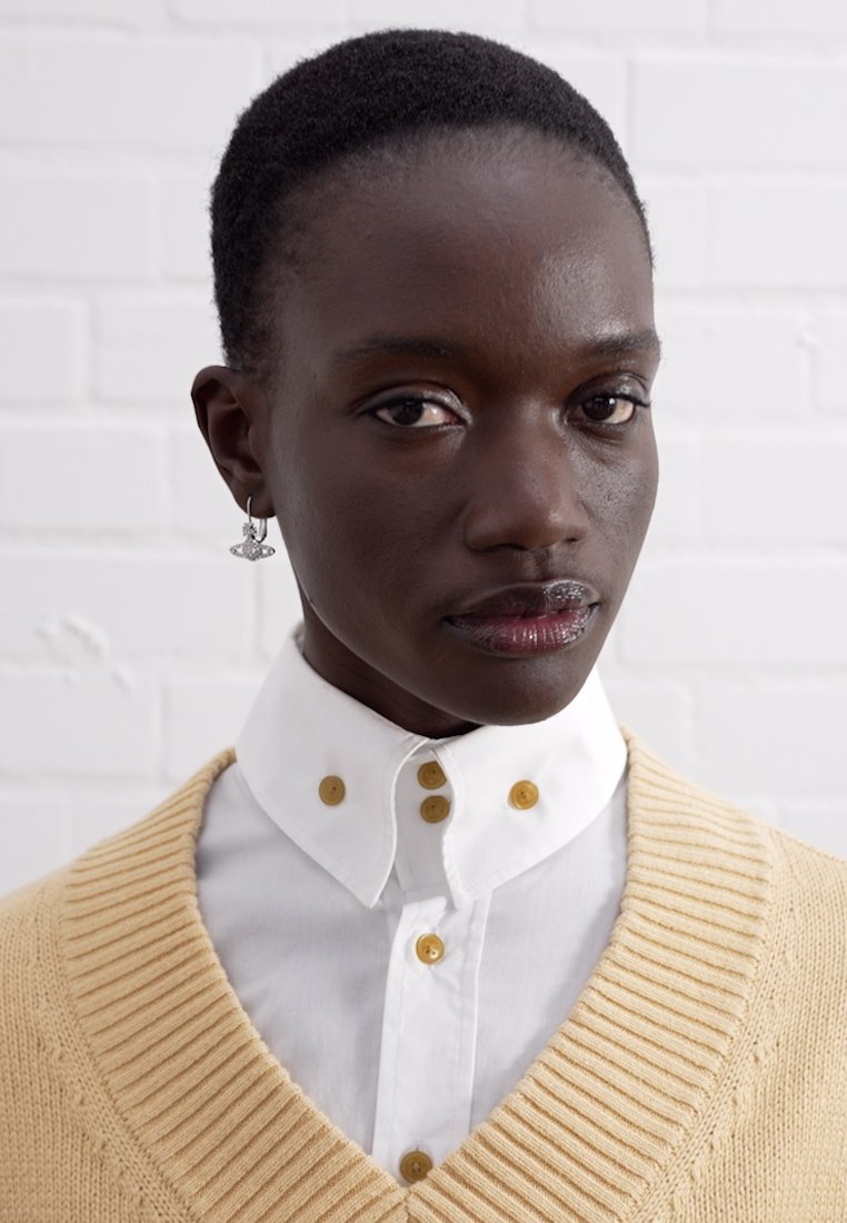 Pull en maille beige clair avec un décolleté en V profond sur une chemise blanche à col. Des boutons dorés sont visibles sur la chemise et de petites boucles d'oreilles en anneau.