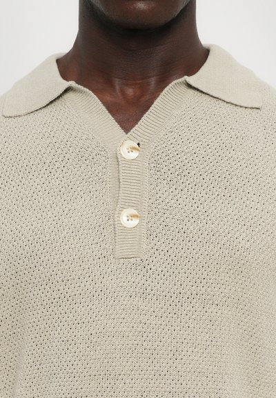 Ανοιχτό μπεζ πλεκτό πόλο πουκάμισο με υφή, V-λαιμό και τρία κουμπιά με κρεμ λευκές, γυαλιστερές λεπτομέρειες.