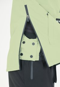 Vihreä vedenpitävä takki, jossa on vetoketjulliset taskut, sisäpuolella napit ja kontrastiväri tummasta kankaasta. Lähikuva muotoilusta ja toimivuudesta.