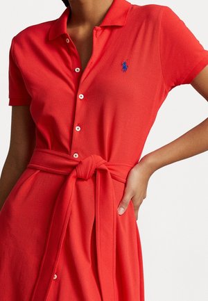 Kobieta w czerwonej sukience polo z guzikami z przodu i paskiem wiązanym w talii, z małym niebieskim haftowanym logo na piersi.