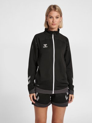 Femme portant une veste de sport noire à fermeture éclair et un short noir avec des motifs chevrons gris, debout devant un fond clair uni.