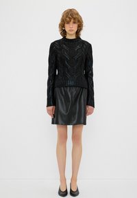Schwarzer gestrickter Pullover mit strukturiertem Muster, kombiniert mit einem schwarzen Leder-Minirock. Das Model trägt schwarze Ballerinas und posiert vor einem neutralen Hintergrund.