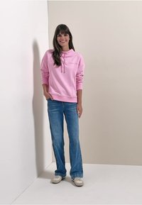 Hoodie di cotone rosa con tasca frontale e coulisse, abbinato a jeans flare azzurri e sneakers beige. Vestibilità semplice e rilassata.