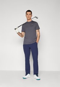Šedé tričko s krátkým rukávem a logem, námořnické kalhoty, bílé golfové boty a bílá rukavice, držící golfovou hůl na neutrálním pozadí.